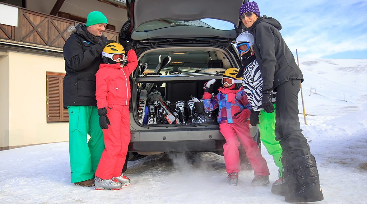 Family of five dressed in ski gear gathered around boot of car filled with ski equipment